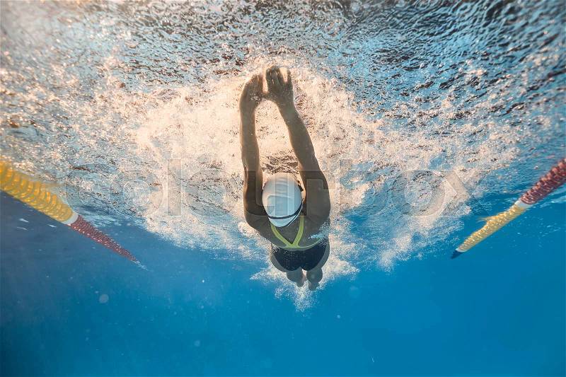 Female swimmer swims in back crawl style underwater in the swimming pool outdoors. She wears a black-lime swimsuit, a white swim cap and swim glasses. Sunlight falls from above. Horizontal, stock photo
