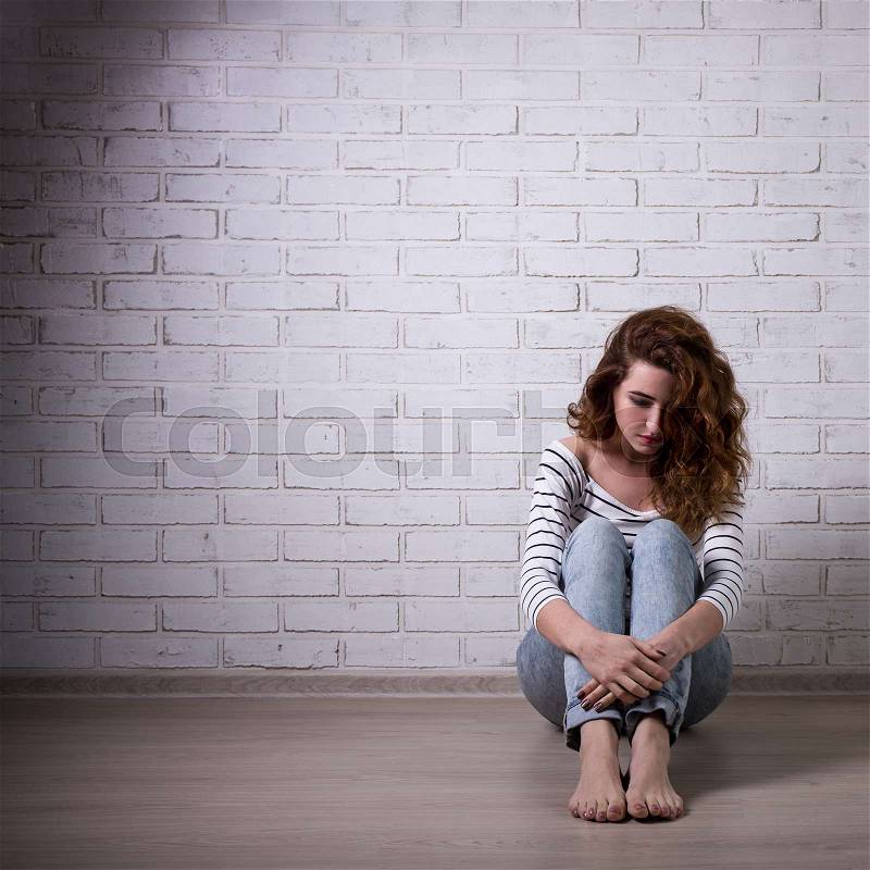 Depression and loneliness - unhappy woman sitting on the floor over white brick wall, stock photo