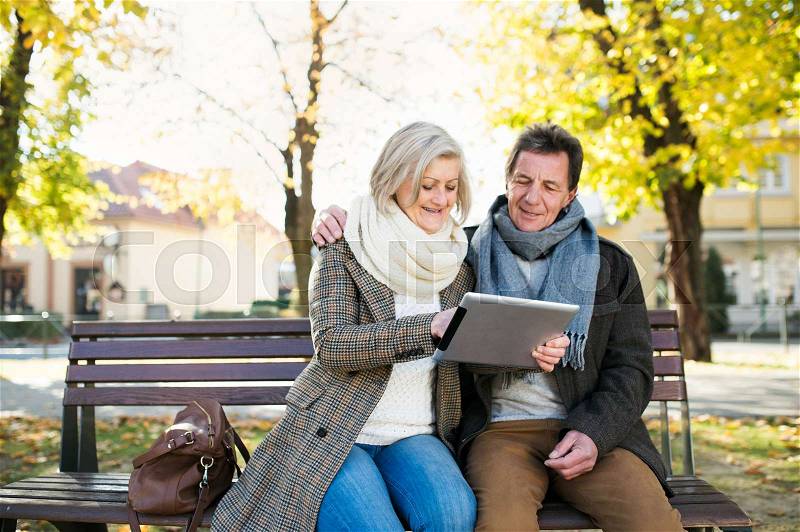 Senior couple with tablet sitting on bench. Sunny autumn park, stock photo