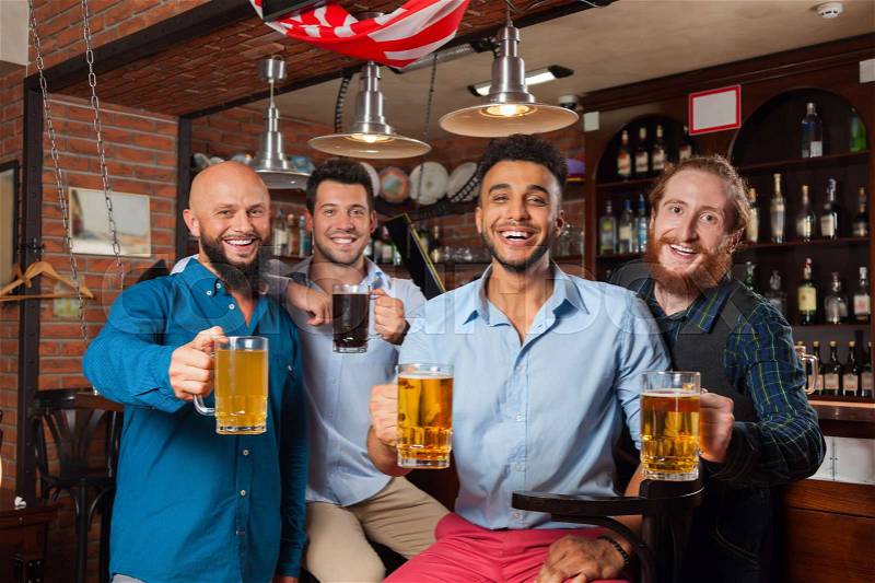 Man Group In Bar Clink Glasses Toasting, Drinking Beer Hold Mugs, Mix Race Cheerful Friends Wear Shirts Meeting Pub Communicate Talking, stock photo