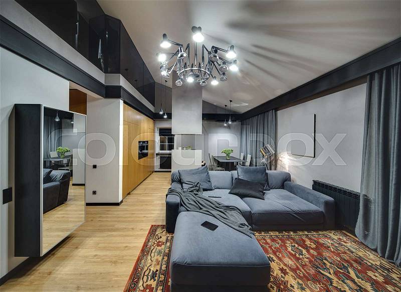 Modern style room with glowing chandelier and lamps. On the floor there is parquet with red carpet. There is mirror, wooden cupboard, kitchen island, table with chairs, blue sofa with pillows, stock photo