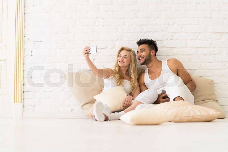 Young Couple Sit On Pillows, Girl Take Selfie Photo Hold Cell Smart Phone, Happy Smile Hispanic Man And Woman Hug Lovers Over White Brick Wall, stock photo