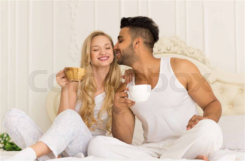 Young Couple Drink Coffee Sitting In Bed, Happy Smile Young Hispanic Man Kisses Woman Lovers Hold Cups Bedroom, stock photo