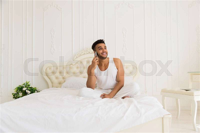 Handsome Hispanic Man Lying In Bed Speak Cell Smart Phone, Young Guy Happy Smile Look Up To Copy Space Morning Bedroom, stock photo