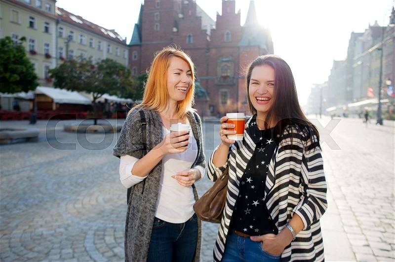 Morning. Nice young women standing on the square of the beautiful European city. They are holding paper cups with coffee and smile. They are surrounded with fine architecture, stock photo