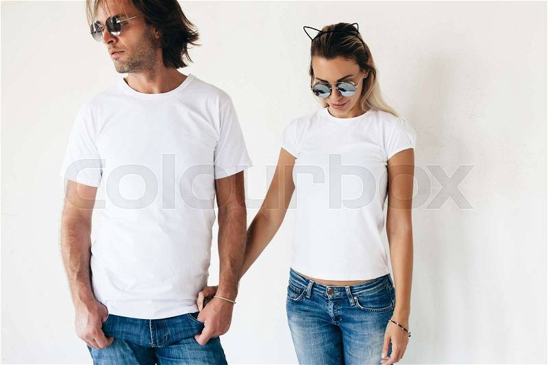 Two hipster models man and woman wearing blanc t-shirt, jeans and sunglasses posing against white wall, toned photo, front tshirt mockup for couple, stock photo