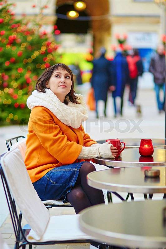 Cheerful young woman in orange coat in Paris decorated for Christmas, drinking coffee, tea or hot chocolate in an outdoor cafe, stock photo