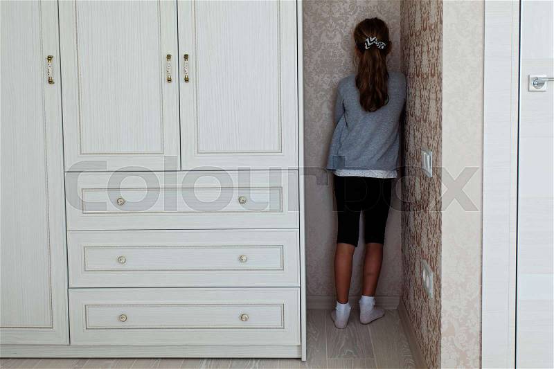 Little girl standing in the corner of her room behind the cupboard , stock photo