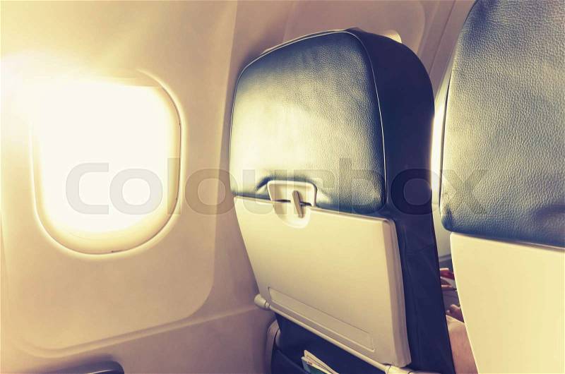 Passenger plane interior fragment. Main cabin chairs with folding tables and glowing porthole with natural sunlight lens flare. Vintage tonal correction filter, warm old style photo effect, stock photo