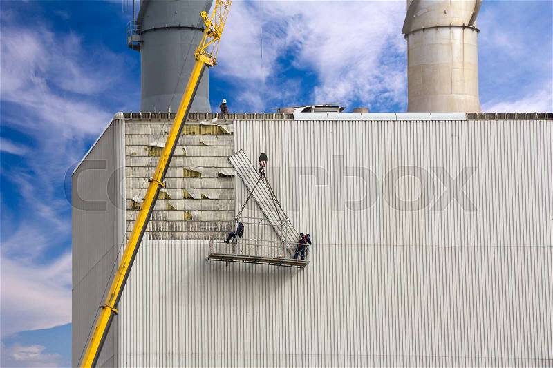 Workers hanging on the crane and repairing wall paneling on heating plant, stock photo