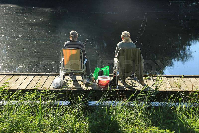 The fishing ladies sitting on the folding chair on the wooden platform and having fun during fishing in the lake in the park in the summer, stock photo