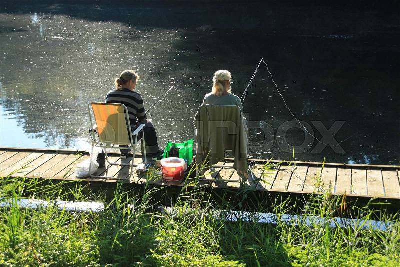 The fishing ladies sitting on the folding chair on the wooden platform and having fun during fishing in the lake in the park in the summer, stock photo