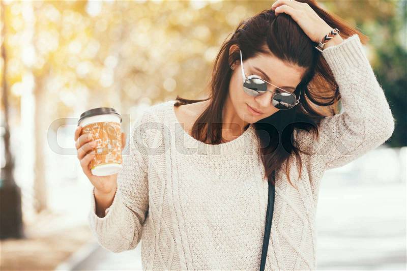 Young woman wearing woolen sweater walking in the autumn city street and drinking take away coffee in paper cup. Breakfast on the go, stock photo