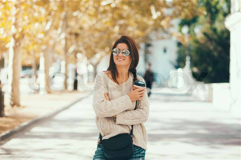 Young woman wearing woolen sweater walking in the autumn city street and drinking take away coffee in paper cup. Breakfast on the go, stock photo