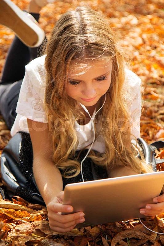 Pretty teenager girl is lying down in autumn park with yellow leaves with digital tablet, stock photo