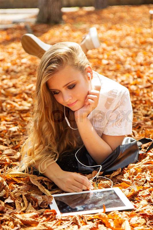 Pretty teenager girl with long hair is lying down in autumn park with yellow leaves with digital tablet, stock photo