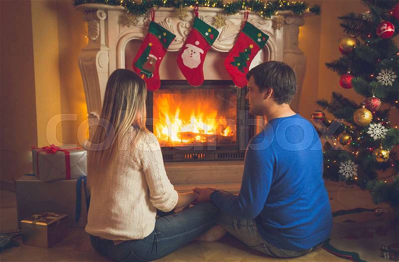 Toned image of young couple in love sitting by the fireplace decorated or Christmas and looking at fire, stock photo
