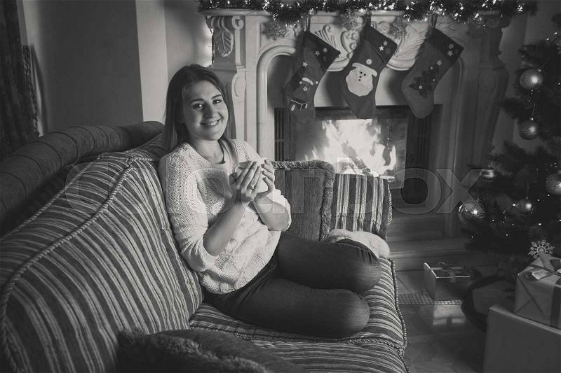 Black and white portrait of happy smiling woman drinking tea on sofa at fireplace decorated for Christmas, stock photo