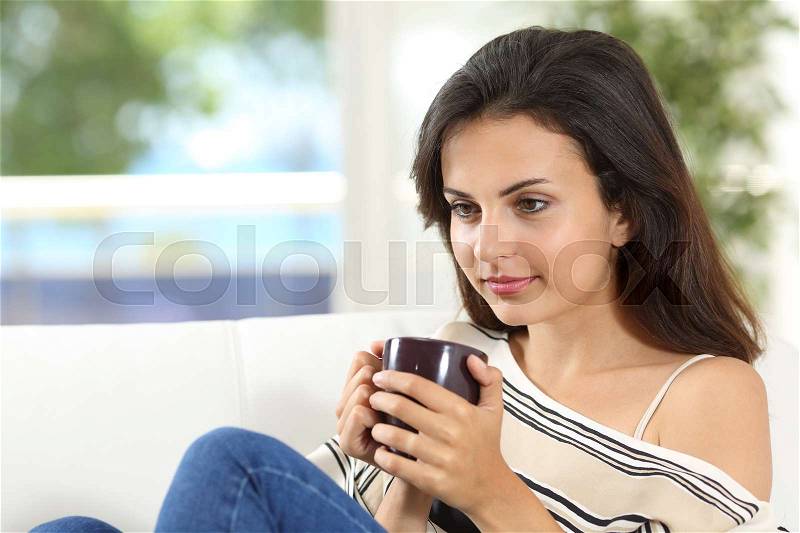 Portrait of a pensive woman thinking with a cup of coffee sitting on a couch at home, stock photo