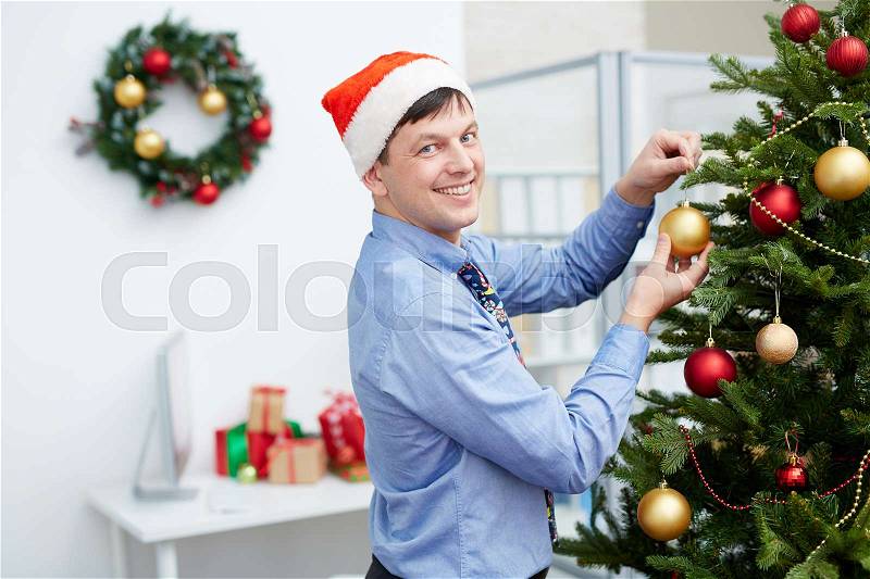 Young smiling businessman in Santa cap hanging bubbles on firtree branches, stock photo