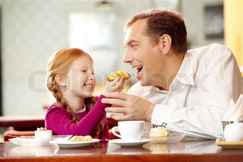 Little daughter giving pastry to her father in cafe, stock photo