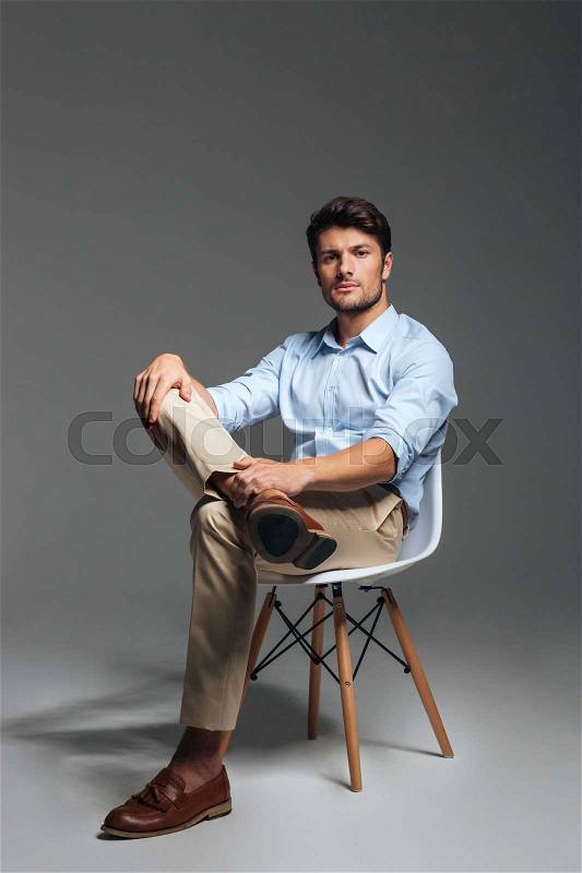 Handsome young confident man sitting on the chair and looking at camera over grey background, stock photo