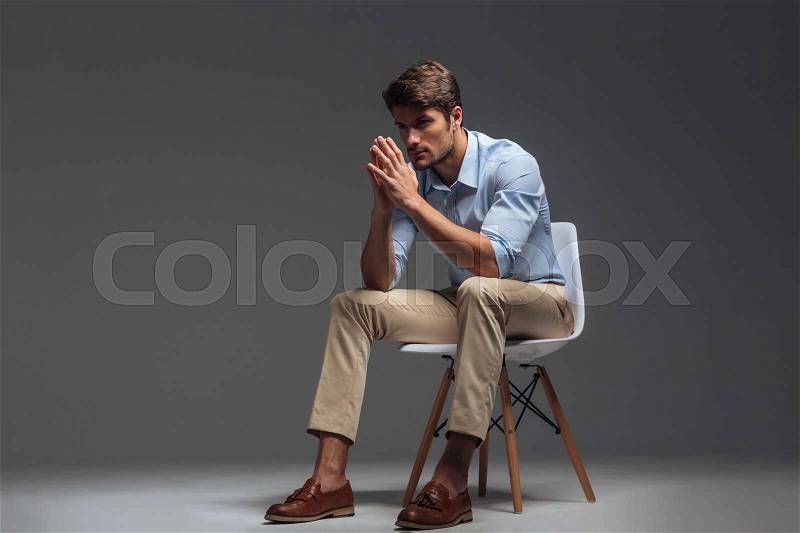 Thoughtful handsome young man sitting on the chair and looking away over grey background, stock photo