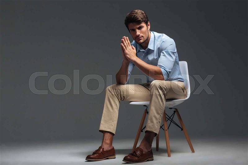 Thoughtful handsome young man sitting on the chair and looking camera over grey background, stock photo