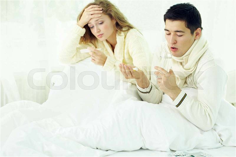 Ill couple sitting in bed, taking temperature and taking medicine, stock photo