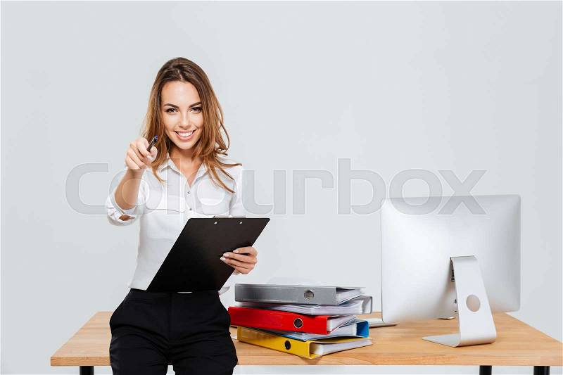 Cheerful young businesswoman holding folder and pointing pen at camera at the office, stock photo