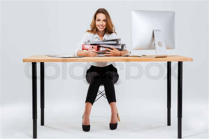 Full length portrait of a happy businesswoman holding folders while sitting at the office desk isoltaed on the white background, stock photo