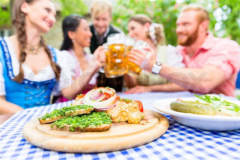Five friends, women and men, sitting in beer garden clinking glasses having Bavarian appetizers on the table, stock photo