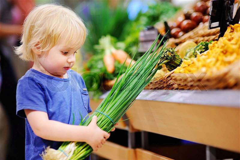 Cute toddler boy in a food store or a supermarket choosing fresh organic green onion. Healthy food for young family with kids, stock photo