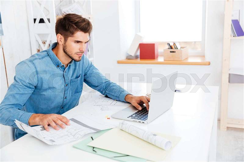 Concentrated young businessman working with documents while sitting with laptop at the office, stock photo