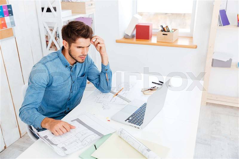 Concentrated young businessman working with documents while sitting with laptop at the office, stock photo