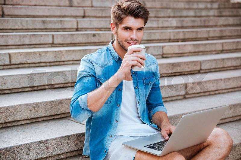 Happy casual man drinking take away coffee and holding laptop while sitting on the sraircase outdoors, stock photo
