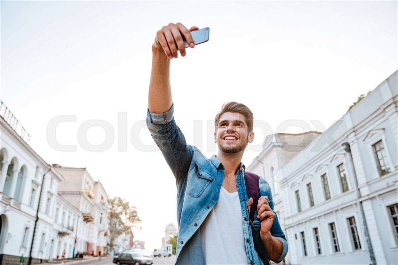 Smiling tourist with backpack making selfies isolated on beautiful modern buildings located in the city centre, stock photo