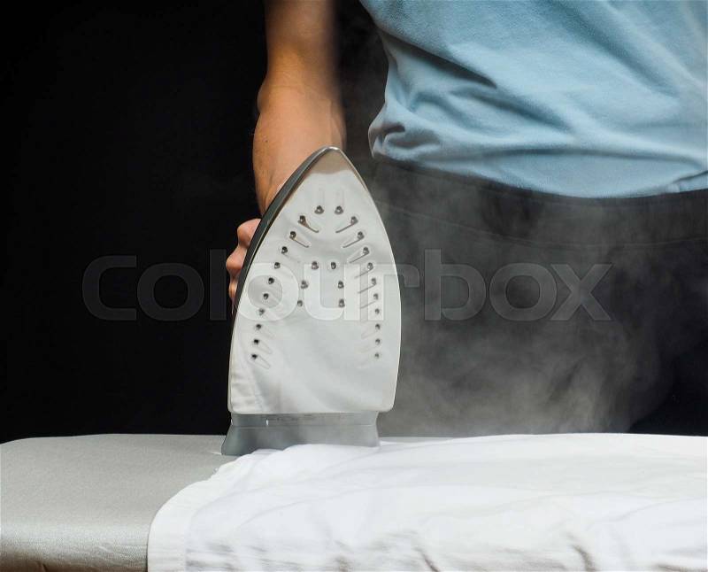 Male person using a steaming hot iron on a white shirt with black buttons, stock photo