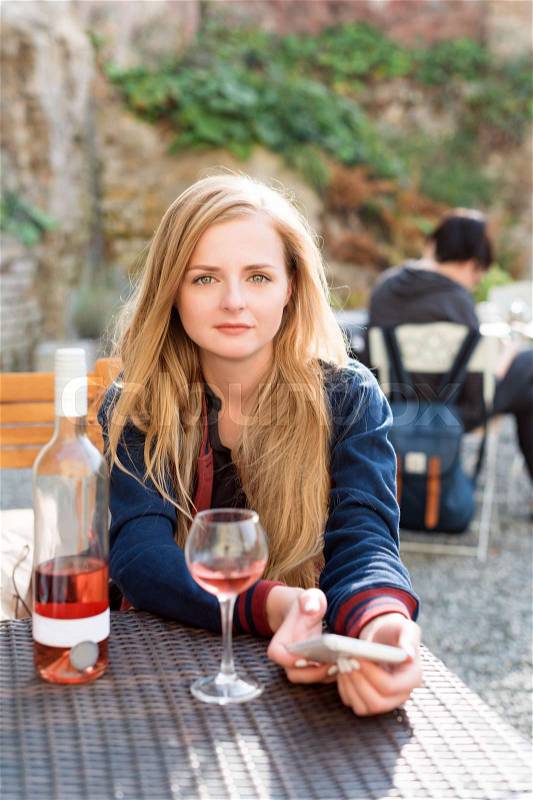 Woman enjoying wine in outdoor city street restaurant or cafe terrace. Portrait of a beautiful wine tasting tourist girl sitting at the table in sidewalk cafe and using her smartphone. Communication, technology, leisure and people concept, stock photo
