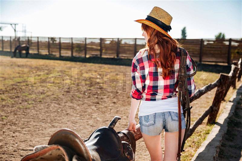 Back view of a young cowgirl in straw hat preparing saddle for a ride, stock photo