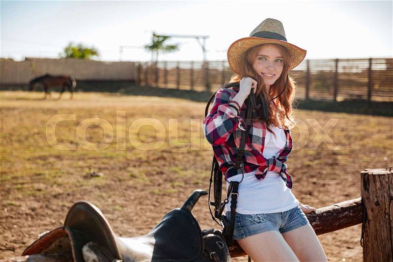 Cheerful cute redhead cowgirl sitting and resting on the ranch fence, stock photo