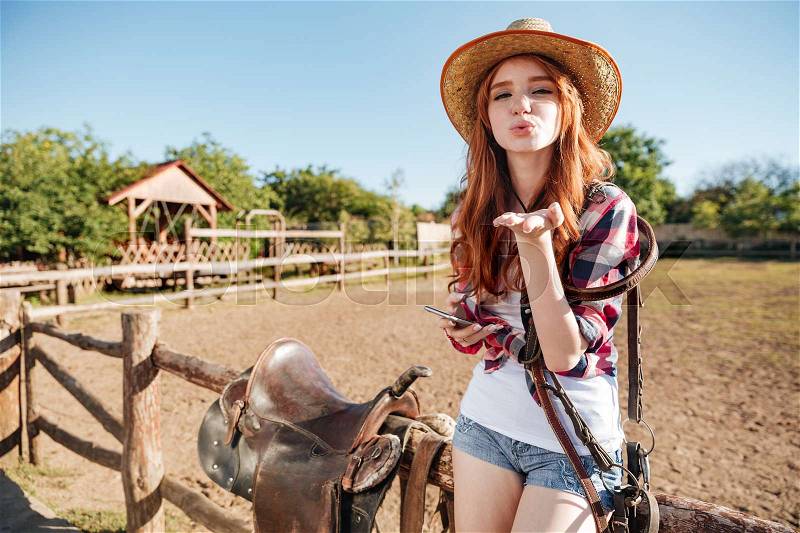 Pretty redhead cowgirl in straw hat sending air kiss while sitting on the fence, stock photo