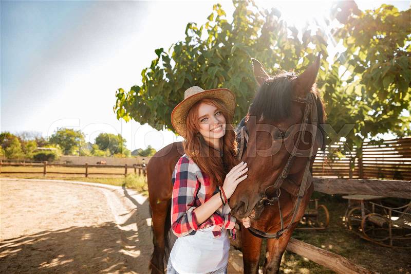 Cheerful beautiful young woman cowgirl standing with her horse in village, stock photo