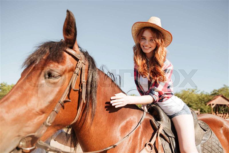 Happy pretty young redhead cowgirl riding horse on ranch, stock photo