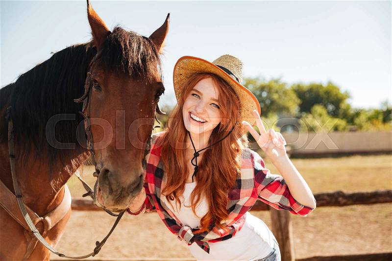 Happy lovely young woman cowgirl standing with horse and showing peace sign, stock photo