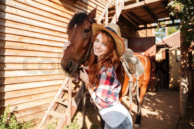 Smiling woman cowgirl in hat with her horse on farm, stock photo