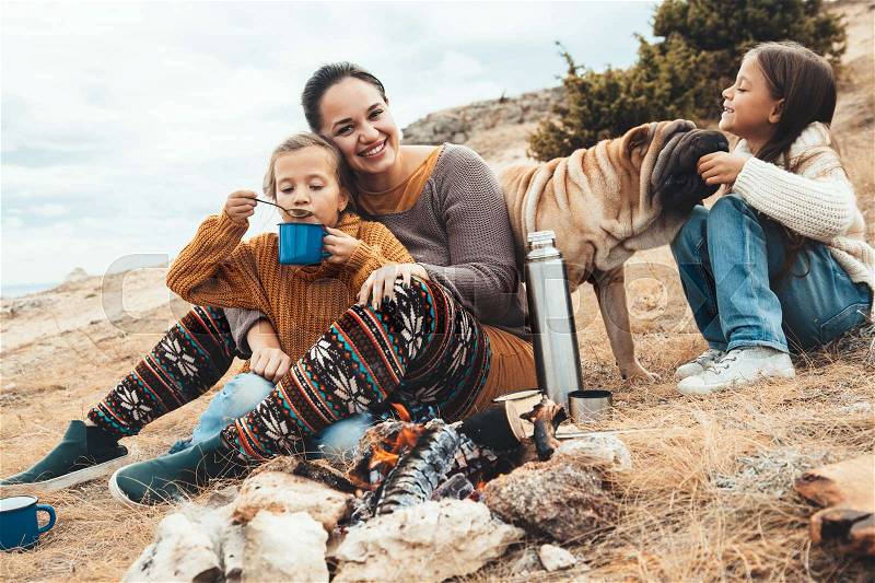 Family with dog sitting by the fire and frying sausages in field. Autumn hike in cold weather. Warming and cooking near flame together, stock photo