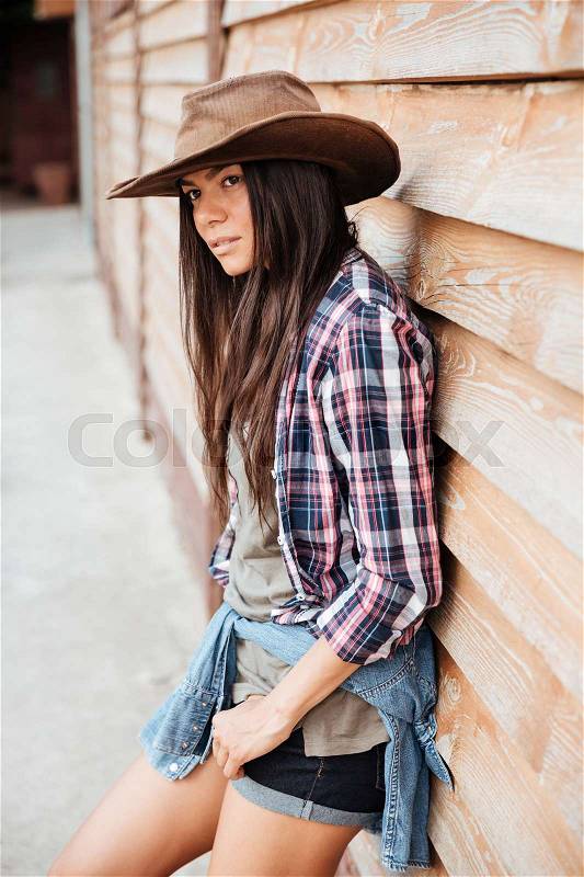 Beautiful young woman cowgirl in hat and plaid shirt standing near wooden house, stock photo
