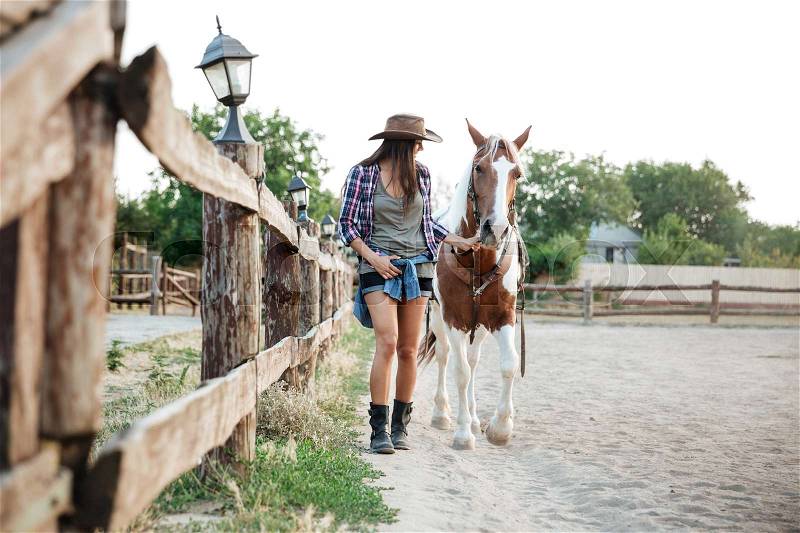 Smiling attractive woman cowgirl holding and walking with her horse on ranch, stock photo