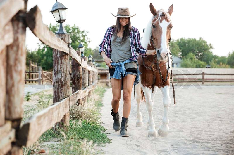 Attractive young woman cowgirl in hat smiling and walking with her horse in village, stock photo